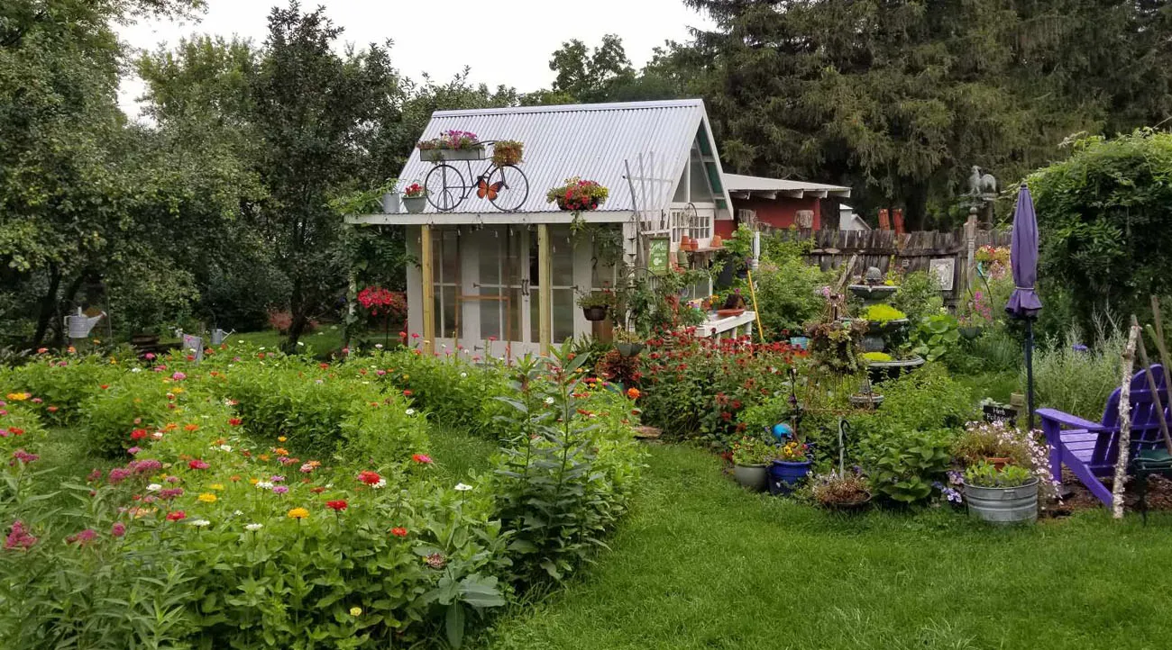 A small structure with a slanted roof sits amongst flower beds. The roof is silver and there is a decorative wire bicycle with flower baskets near the gutter.