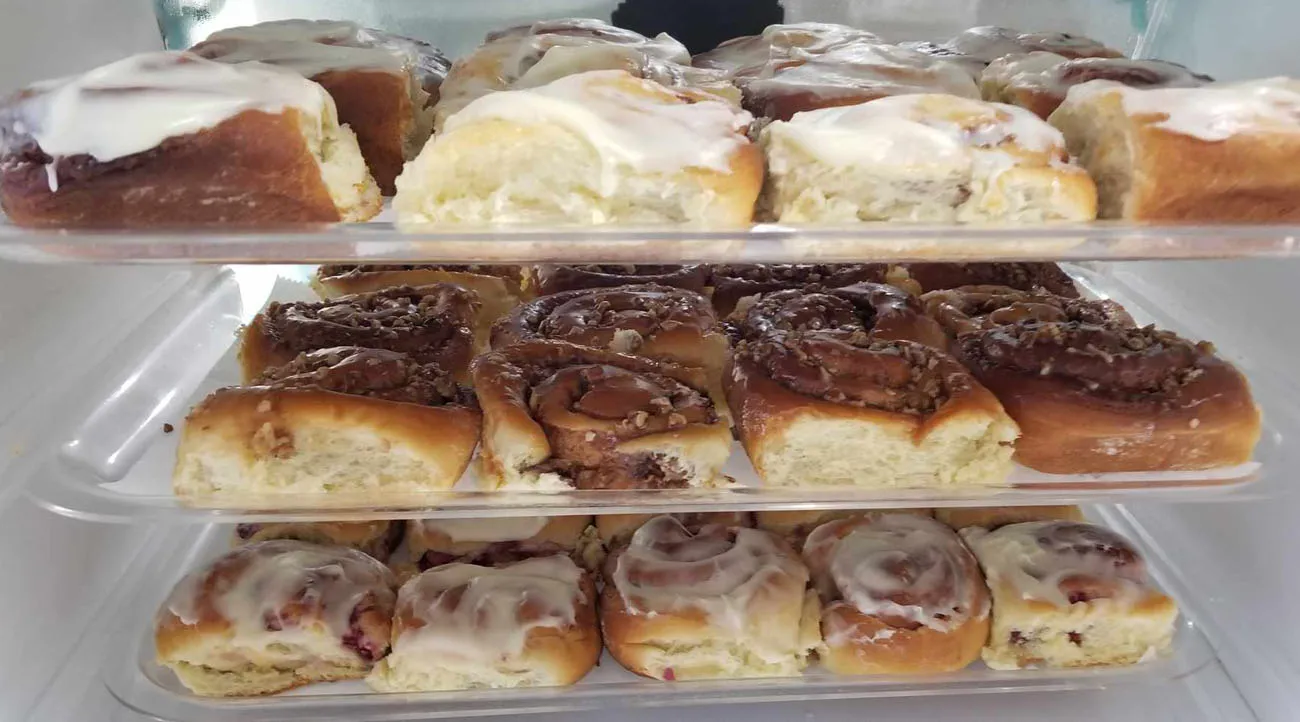 Three rows of pastries on trays are shelved on top of one another. They appear to be variations of cinnamon rolls.