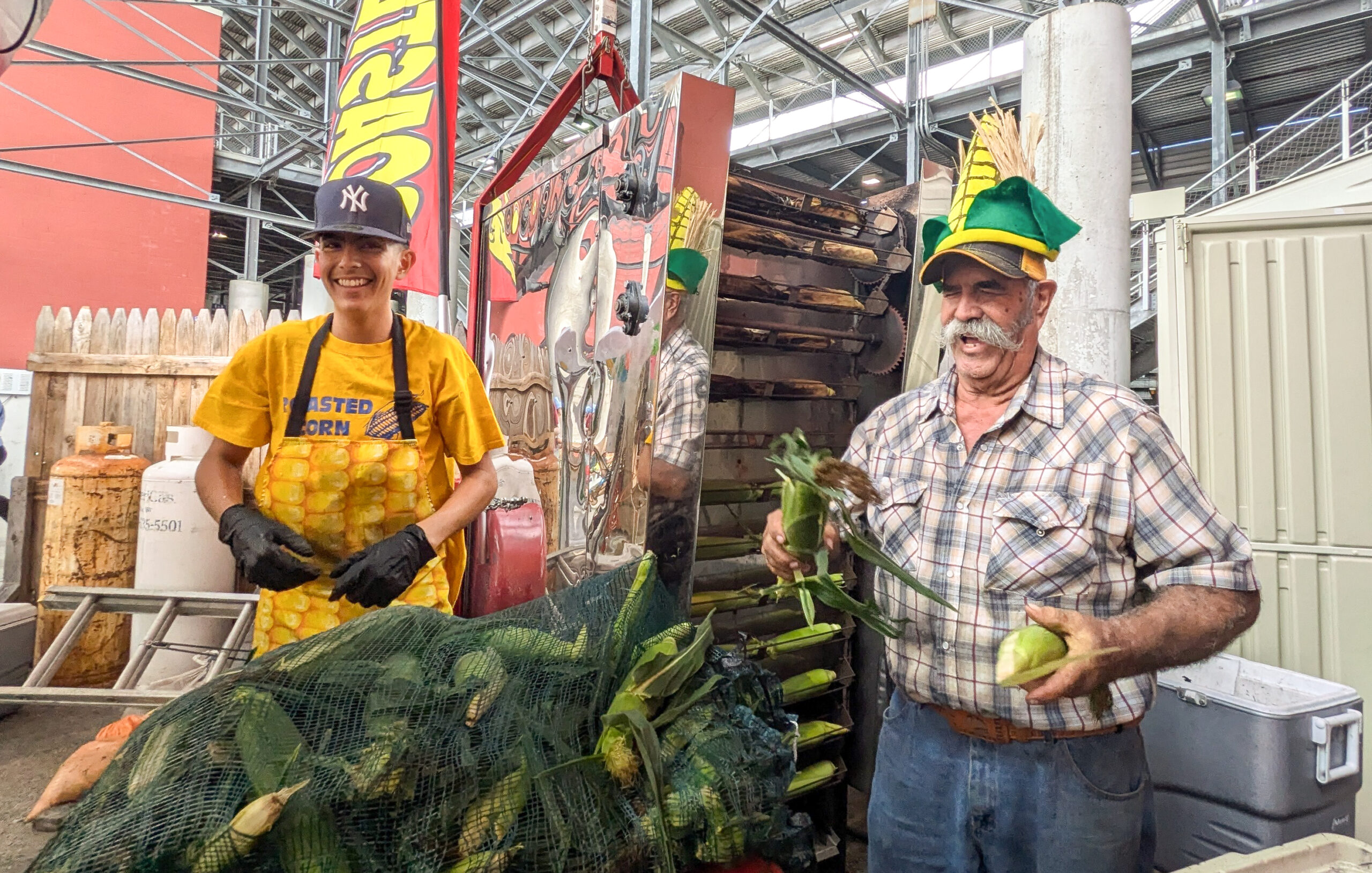 An elderly man with a large, long white mustache laughs. He is wearing a cap that looks like a corn cob while holding cobs in his hands. There is a young adult standing by him wearing a yellow tshirt and an patterned apron with yellow corn kernels.