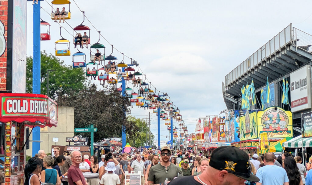 A large crowd walking under a sky glider with people sitting in it.