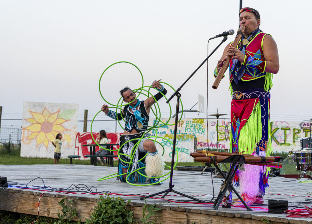 Two people performing on stage, wearing colorful regalia. One of them performs with multiple hoops, while they other plays a flute.