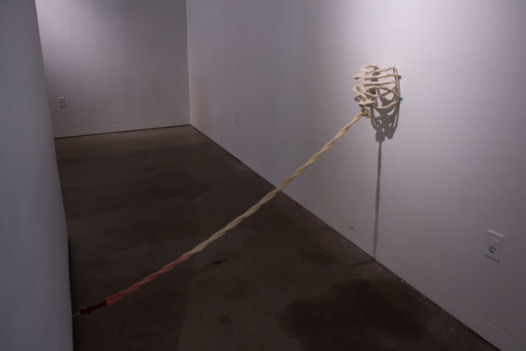 A ceramic ribcage attached to a gallery wall with yarn leading from the breastbone to the floor