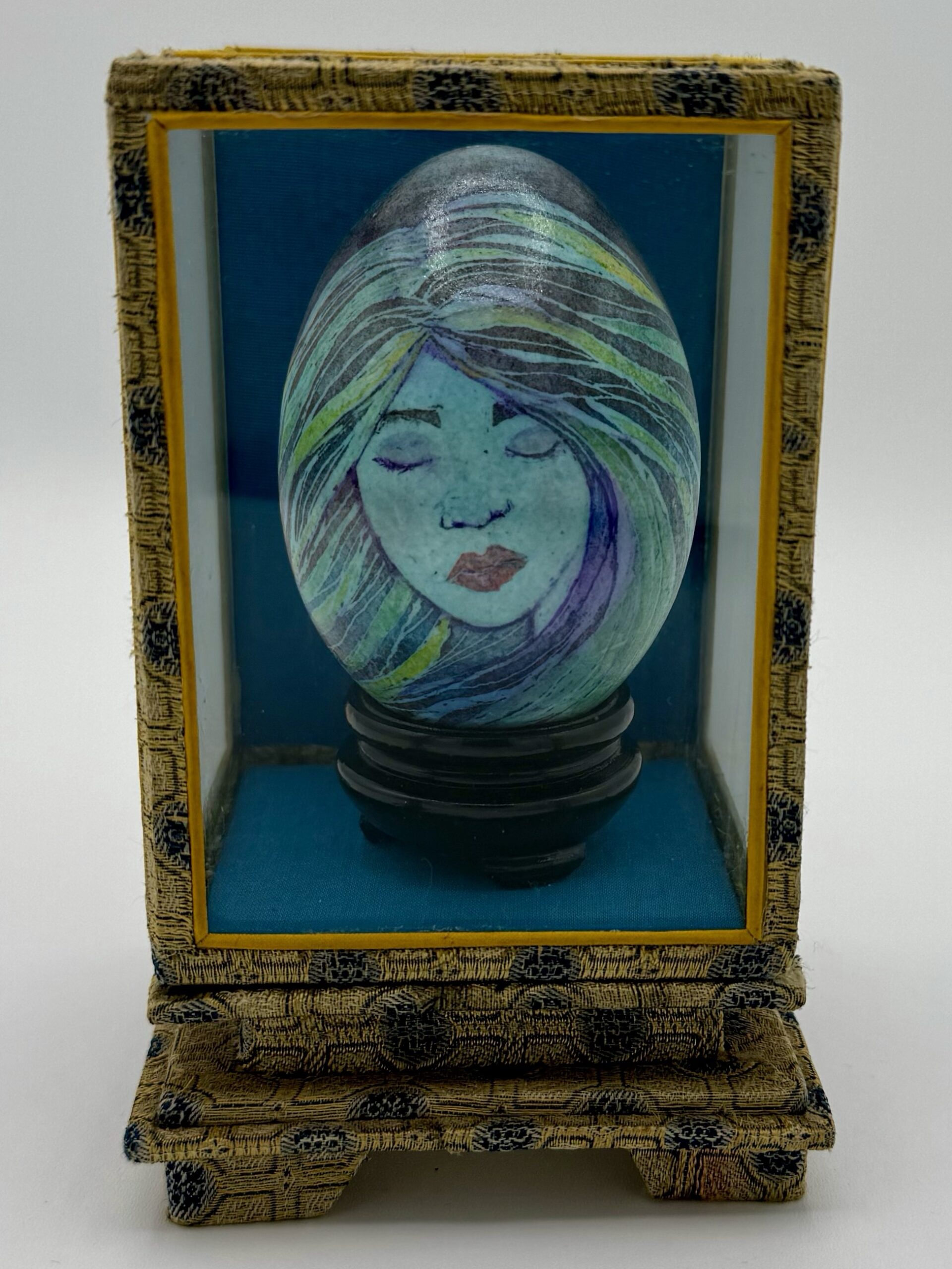 A colorful woman's face on an ostrich egg encased in a glass box decorated with golden fabric.