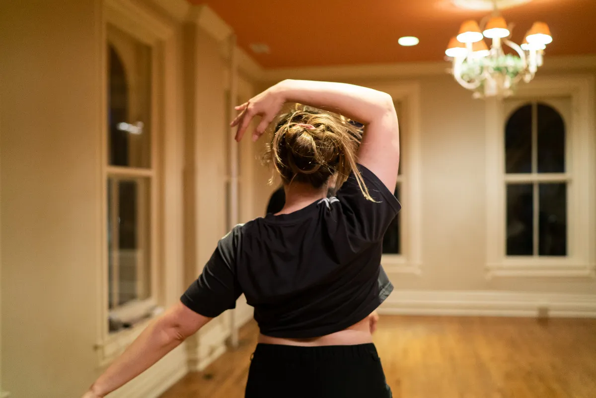 A person in a black t-shirt is facing away from the camera with their right arm raised and curved above their head as their left around is outstretched down and to the left.