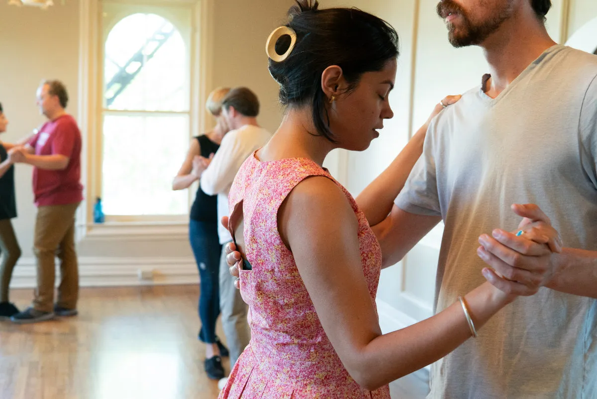 Three couples dance in partner dance formation around a sunny room. The couple in the foreground is a person with dark hair in a yellow clip and a bright pink sundress. Their partner is in a gray t-shirt. The person in the sundress has their eyes closed and mouth slightly open.