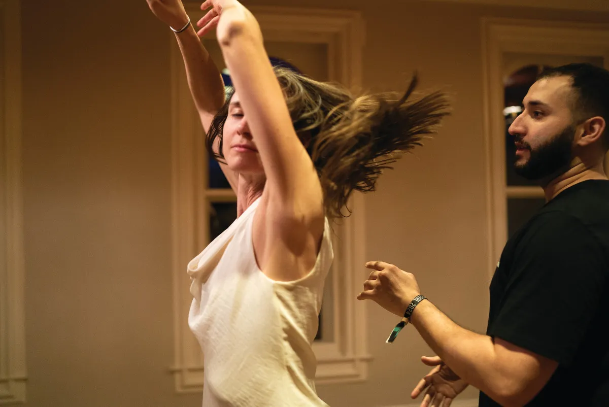 A person in a white tank top spins with their arms up facing away from a person in a black t-shirt as their hair flies in a spiral with the motion.