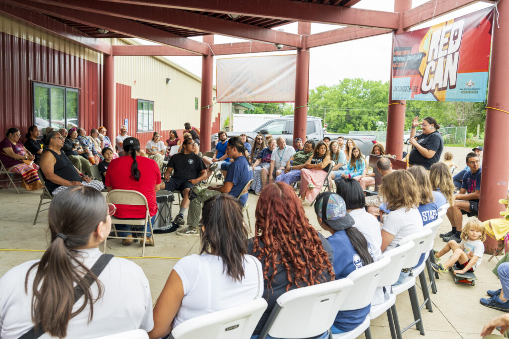 A large group of people sit on chairs arranged in concentric circles under a covered area outdoors. There is a small group of people in the middle with a large drum.