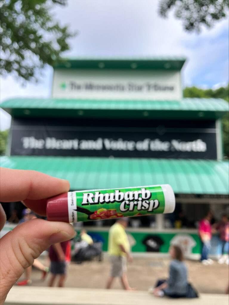 A green lip balm tube reading, "Rhubarb Crisp" in front of a building reading, "The Minnesota Star-Tribune; The Heart and Voice of the North"