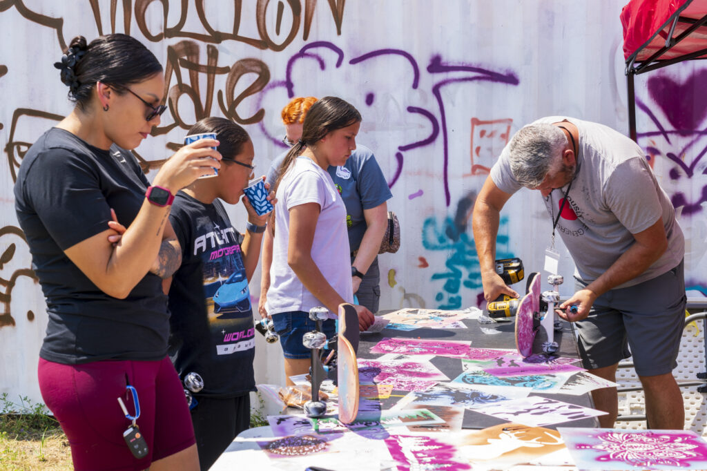 Young people gather around a table with colorful stencils. An adult across from them uses a drill to put wheels on a skateboard.