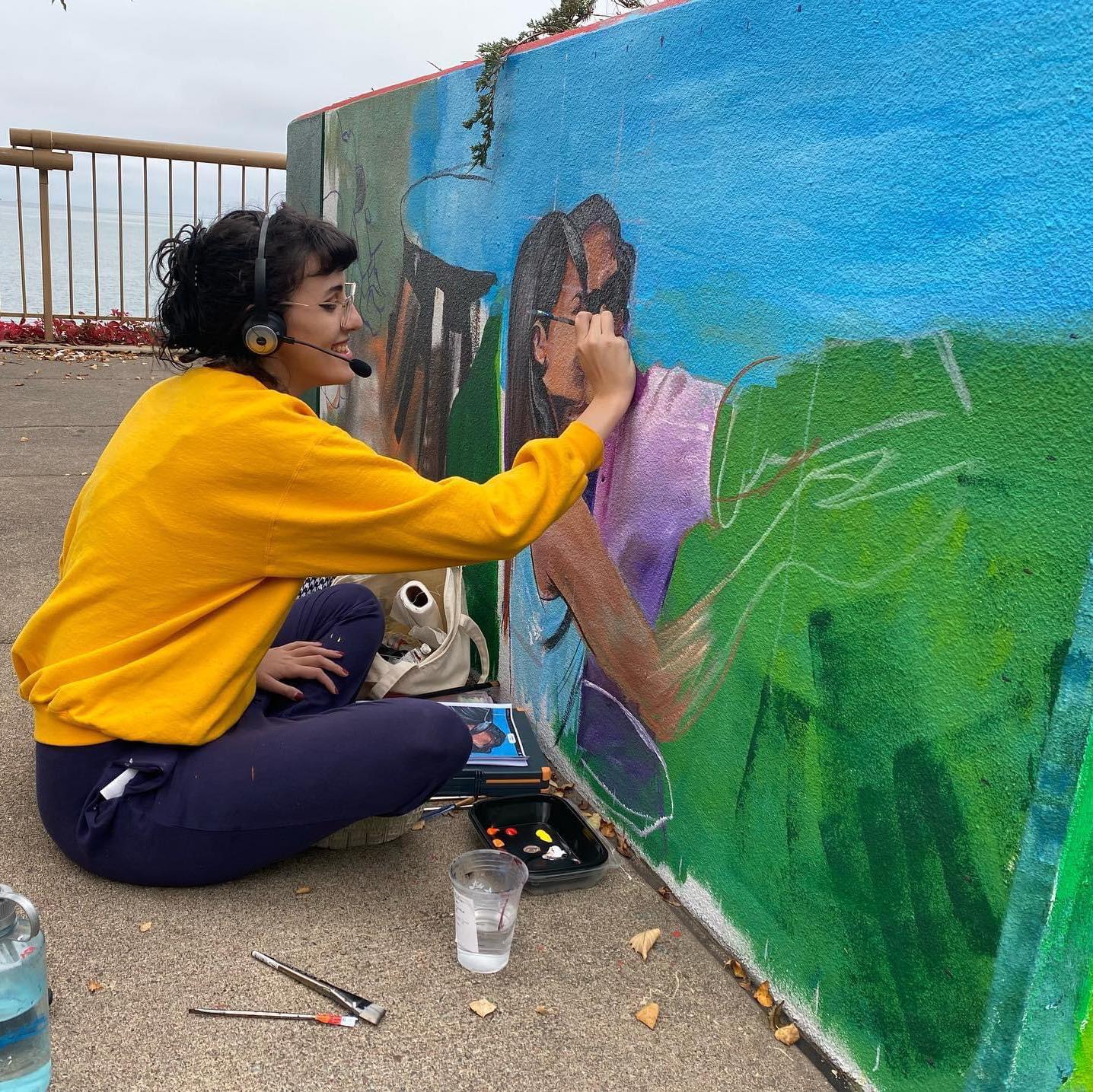 A person in a bright yellow sweatshirt sits on the ground painting a mural on the side of a low wall. In the unfinished scene two people stand close together on bright green grass in front of a bright blue sky. The painter wears a headset with attached microphone.