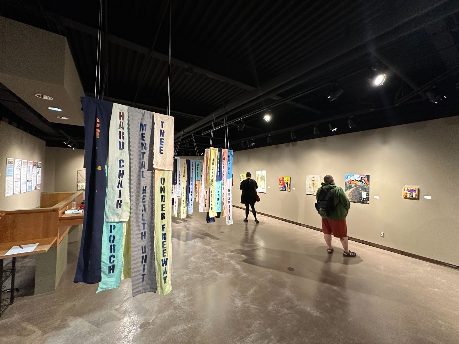 Two people stand looking at paintings on the far side of a gallery wall. In the foreground multicolored fabric hangs together from the ceiling. On the pieces of fabric the words "Hard Chair," "Porch," "Mental Health Unit," "Tree," and "Under Freeway" are printed.