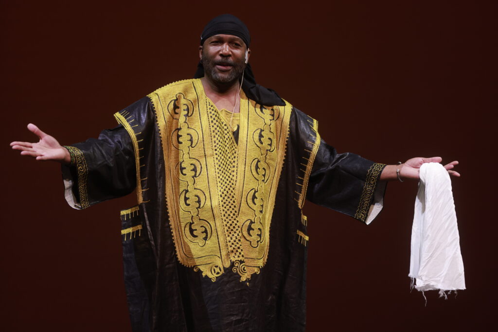 A Black man with a light salt & pepper beard wearing a traditional West African black and gold colored Grand Robe. His arms are reaching out to the sides, and he holds a piece of white fabric in his left hand.