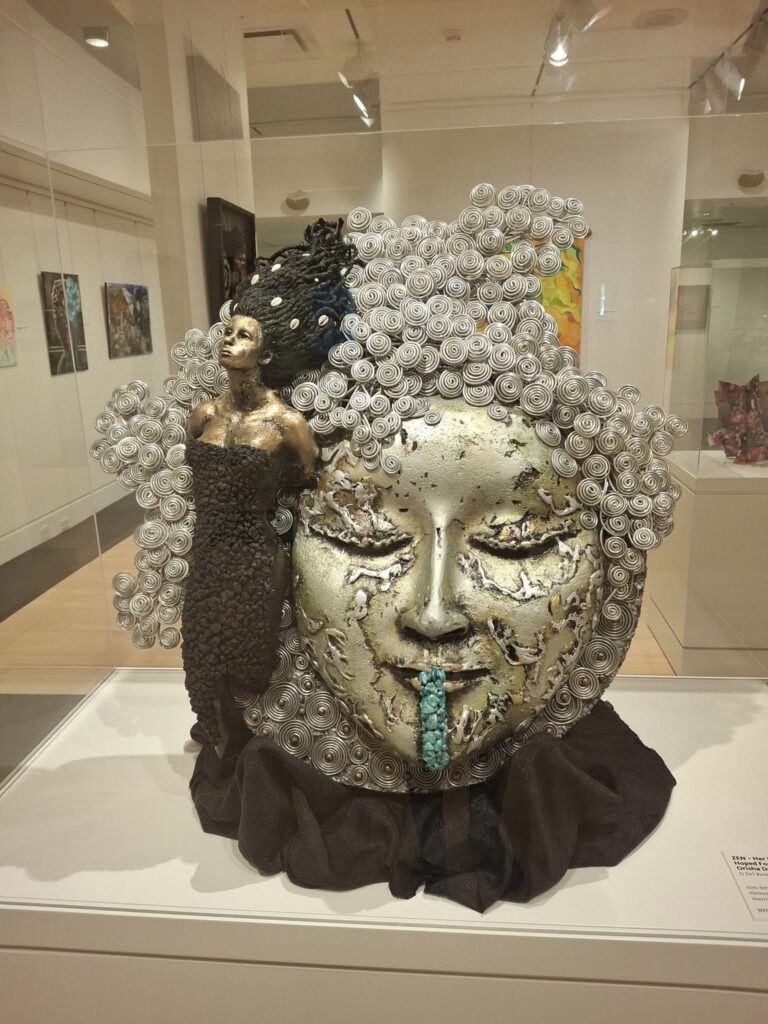 A sculptural work depicting a human figure with long flowing hair against an full-size head with different metal embellishments.