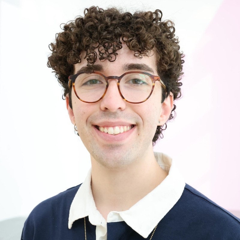 Headshot of a smiling person of light skin tone, with curly dark brown hair, wearing a button up shirt.