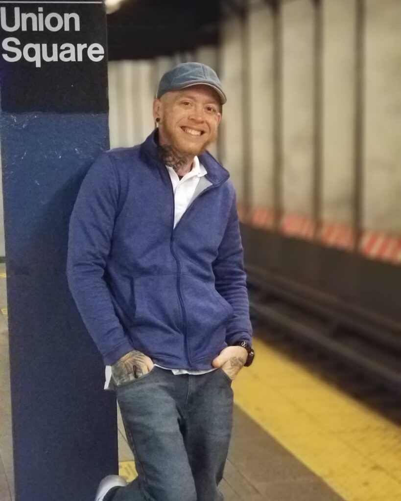 A person wearing a blue jacket poses against a pole in a subway station.