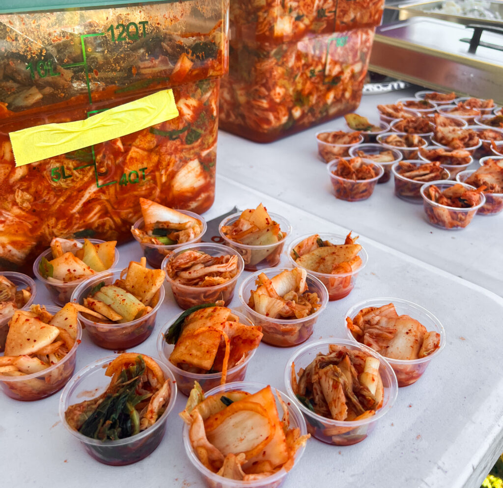 Dozens of small sampler ramekins full of bright red kimchi sitting on a table in front of larger plastic tubs of kimchi.