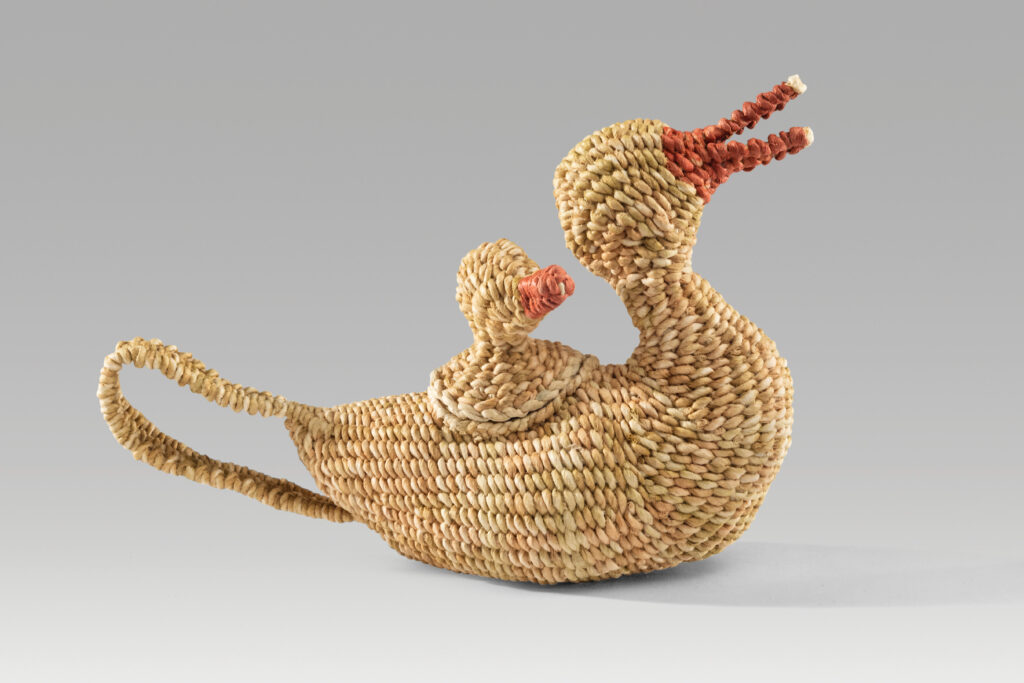 A tan teapot woven out of Korean paper (called hanji) using jiseung (paper basketry) techniques in the shape of a duck with a lid on the back shaped like a duckling, both with reddish pink beaks.