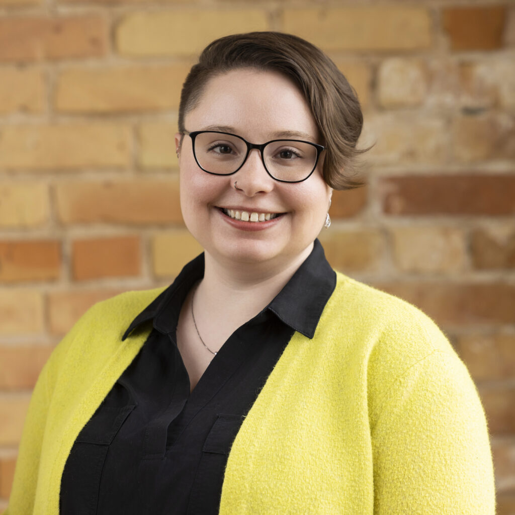 Headshot of a smiling person of light skin tone, with short brown hair pushed to one side, and wearing a black shirt and yellow cardigan