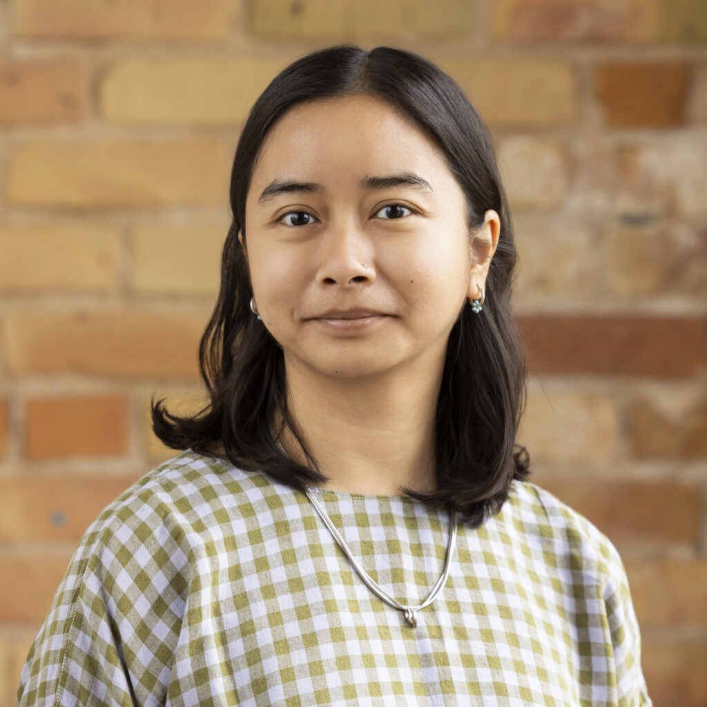 Headshot of a smiling person of medium skin tone, with shoulder length black hair, and wearing a plaid top