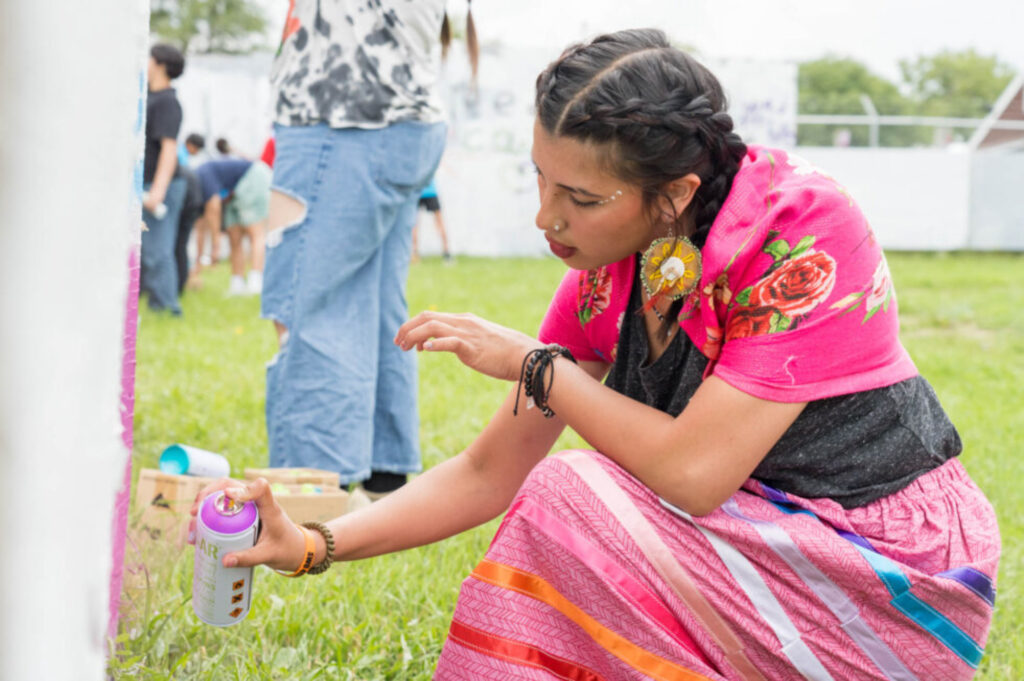 A young Lakota artist in a colorful pink shawl at ribbon skirt paints with a can of pink spray paint