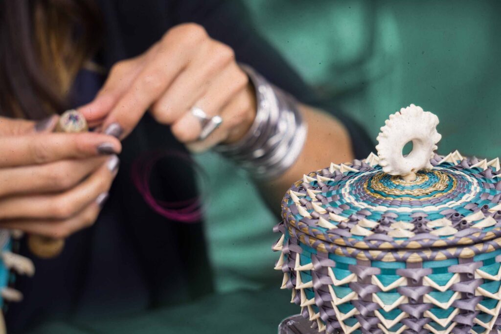 A basketmaker with medium light skin tone weaves a colorful blue and purple basket