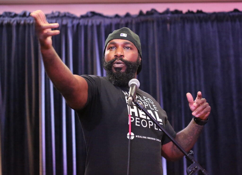 A person with dark skin tone and a backwards baseball cap performs on a microphone with a hand outstretched to the audience