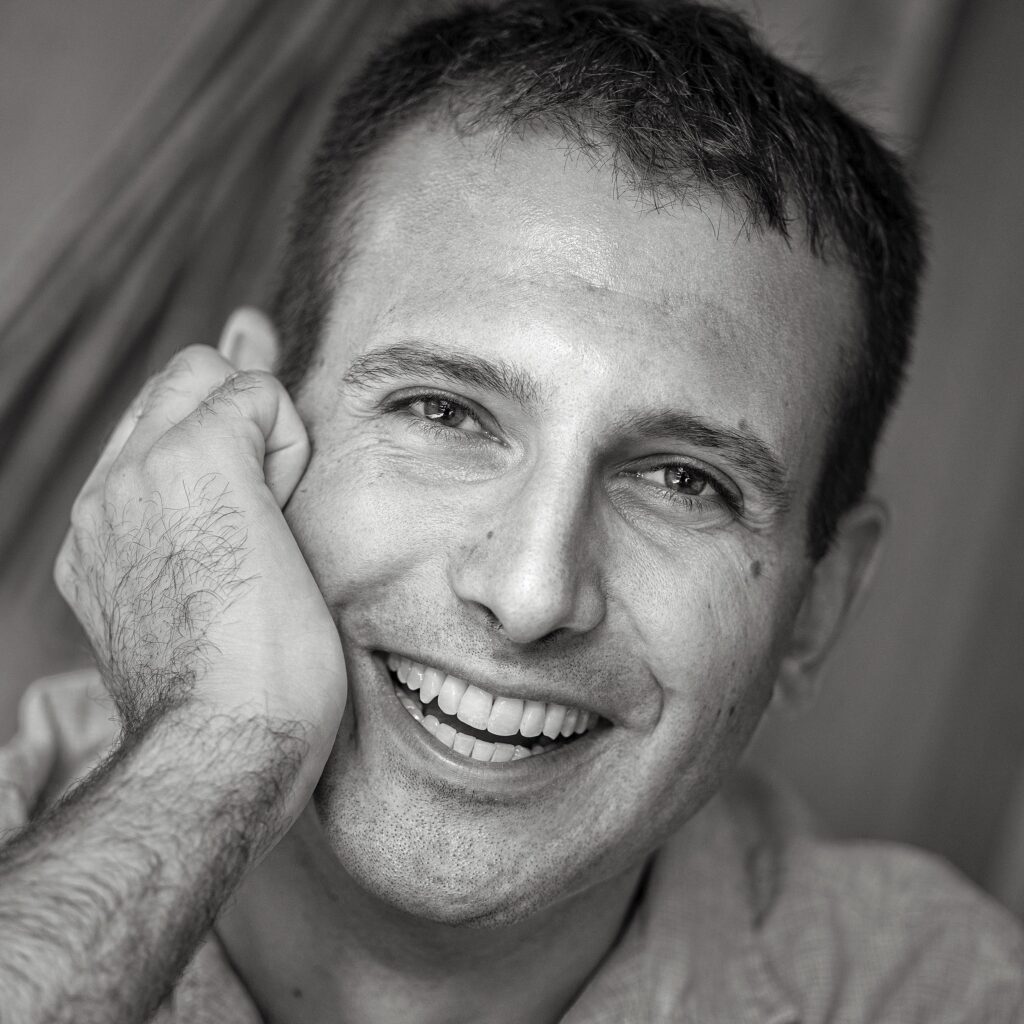 A black and white headshot of a smiling person with short dark hair and light skin tone.