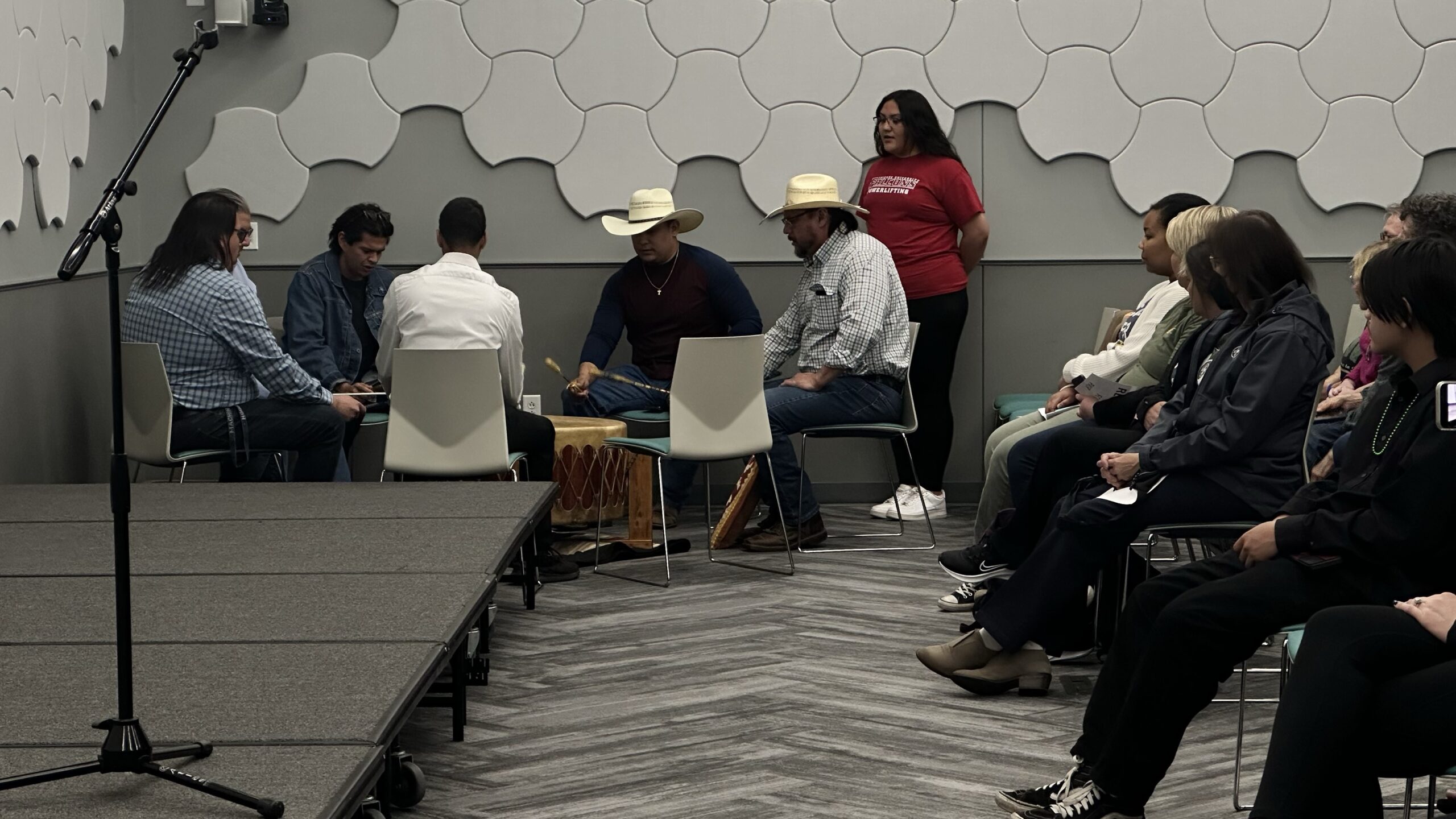 In a grey conference ballroom, a group of Native drummers play a drum in the corner of the room