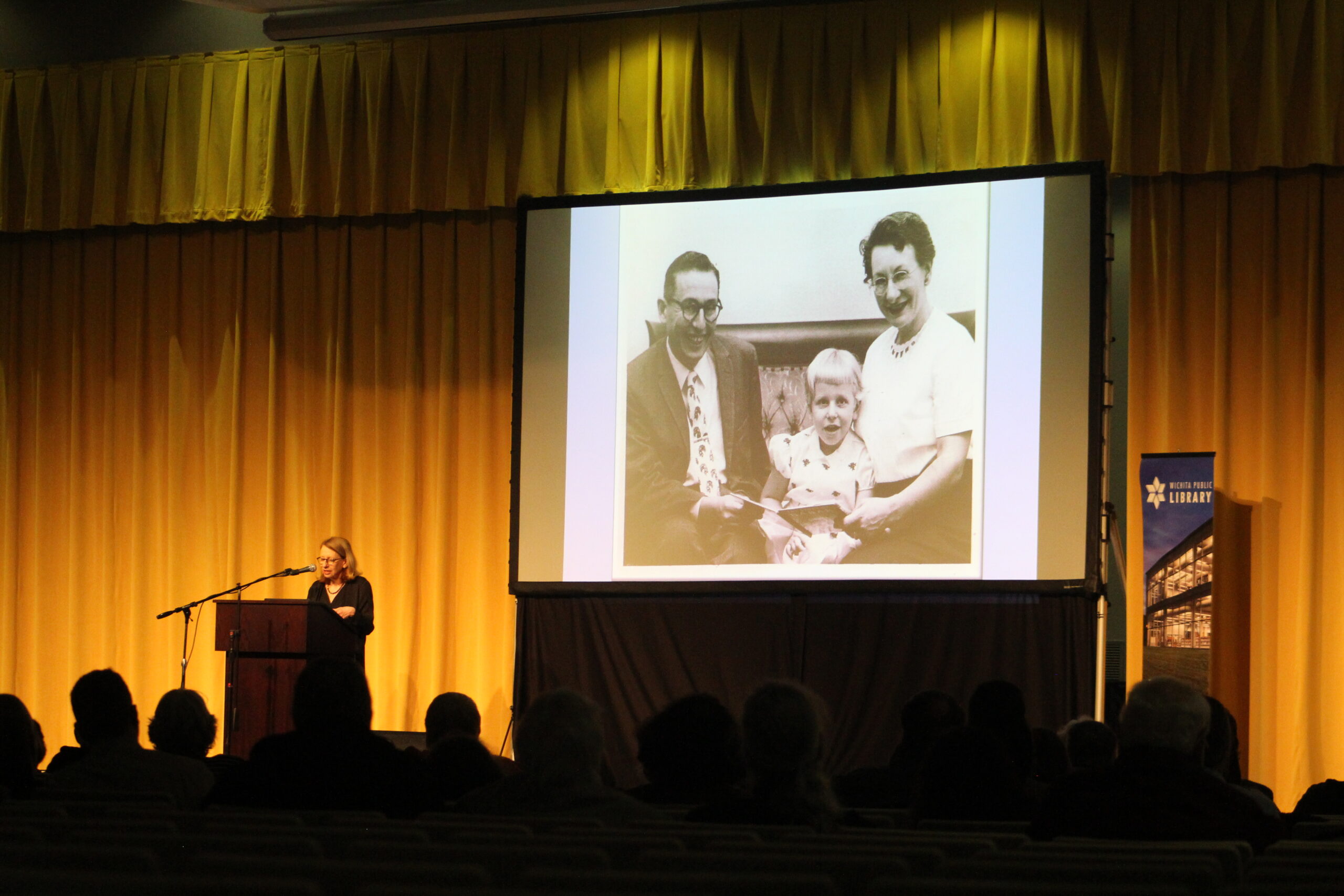 A person speaks to a room of silhouetted figures with a family photo on a screen.