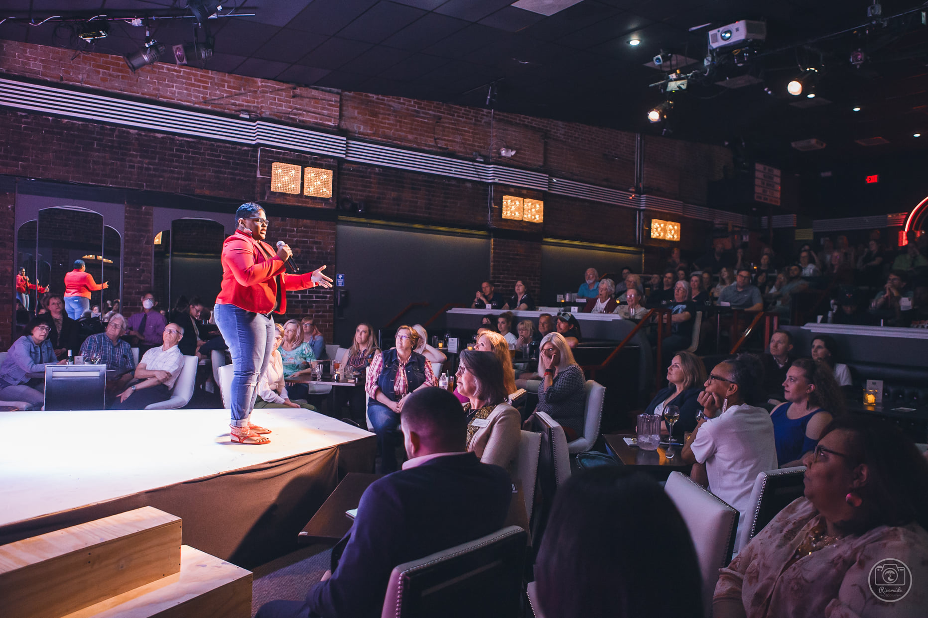 A person with darker skin and a colorful red jacket speaks on a mic in front of a crowd