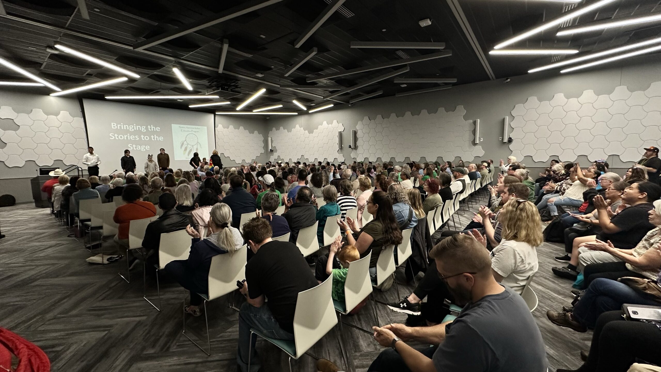 A conference room of people in front of a screen that reads Bringing the Stories to the Stage