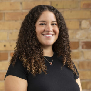 Headshot of a smiling person of medium skin tone, with brown curly hair, and wearing a black shirt.