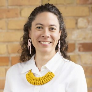 Headshot of a smiling person of light skin tone, with light brown curly hair, and wearing a white shirt and decorative yellow necklace