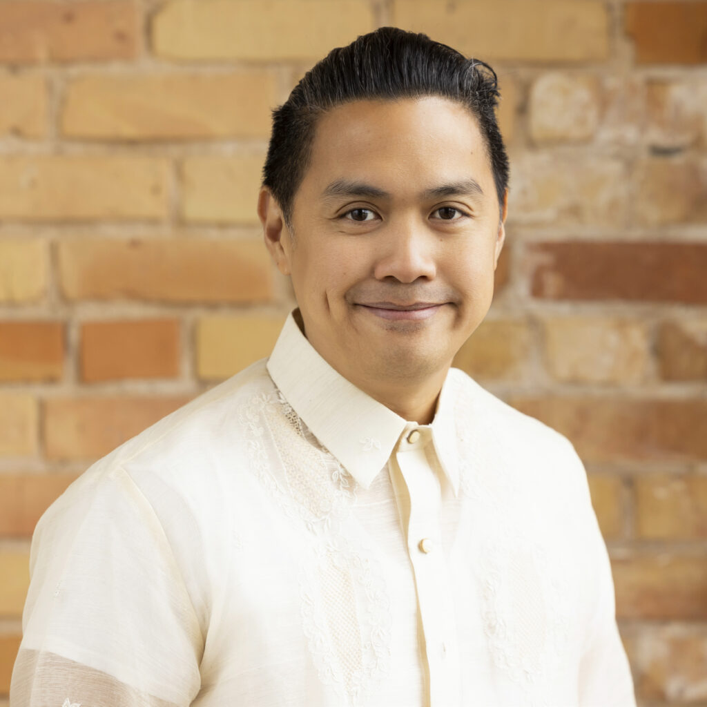 Headshot of a smiling person of medium skin tone, with black short slicked back hair, and wearing a patterned white top.