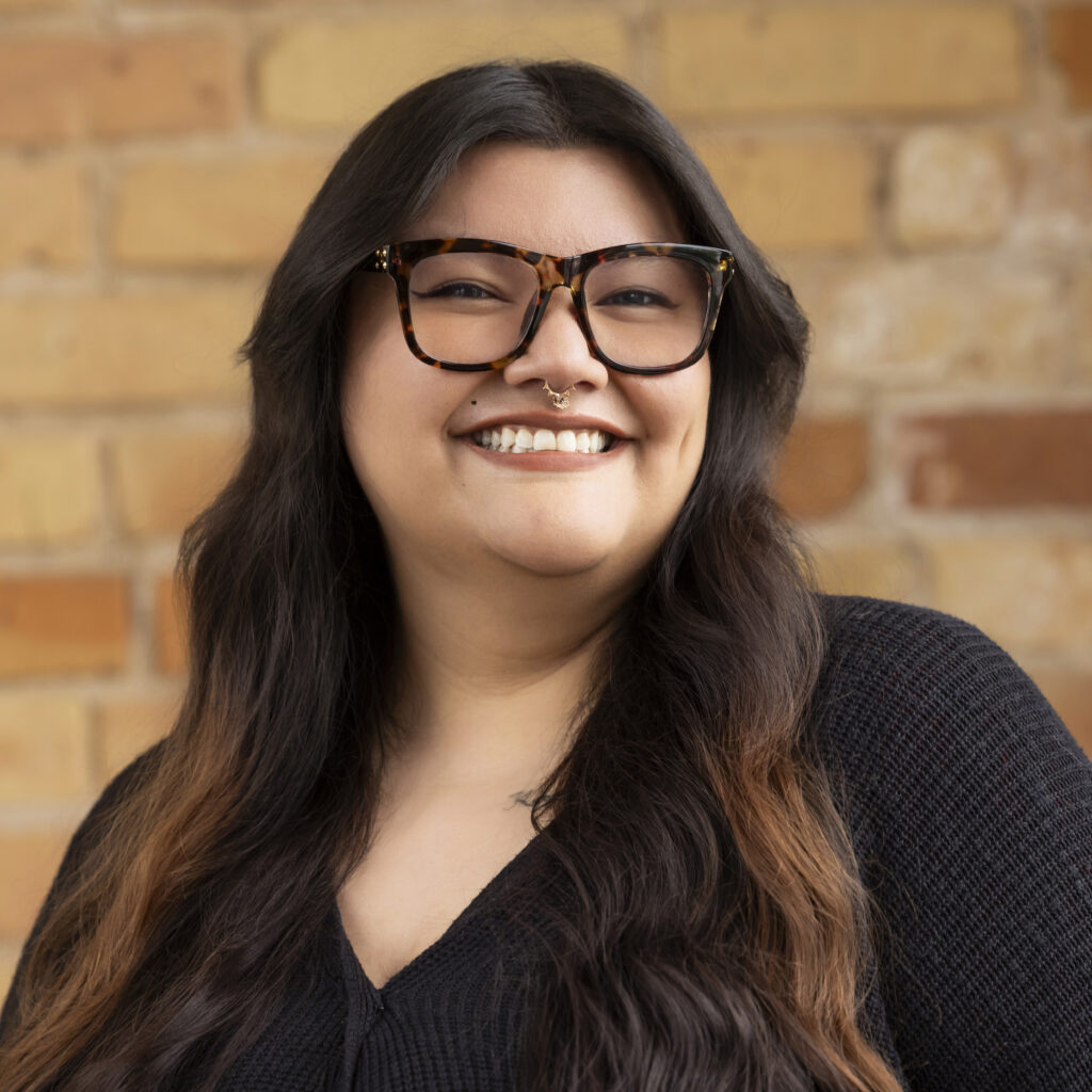 Headshot of a smiling person of medium skin tone, with dark brown hair, glasses, and a nose ring, and wearing a black sweater.