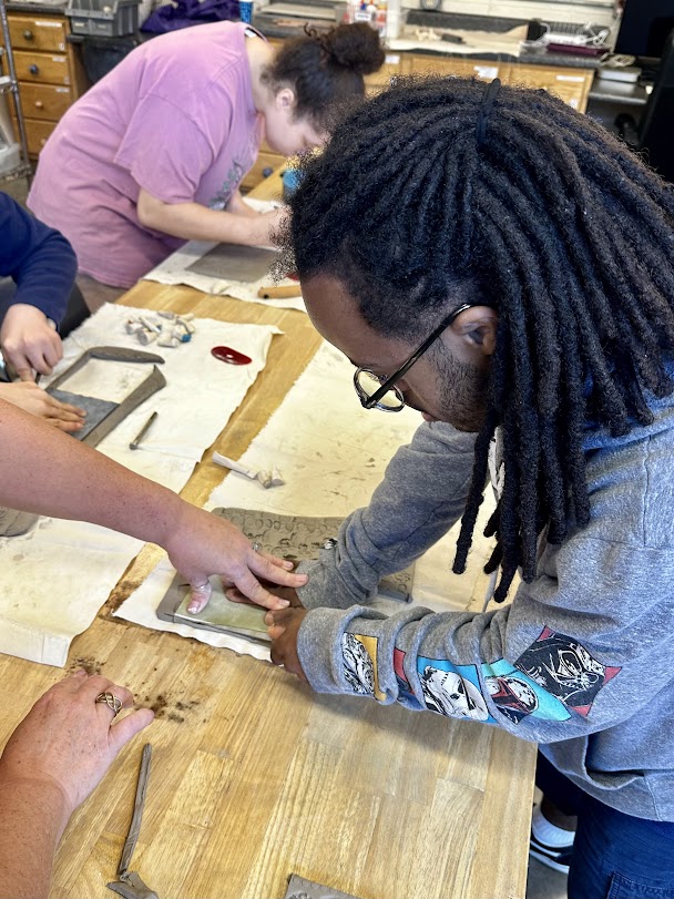 A person with dark skin and deadlocks transfers hand carved stamps on to clay.