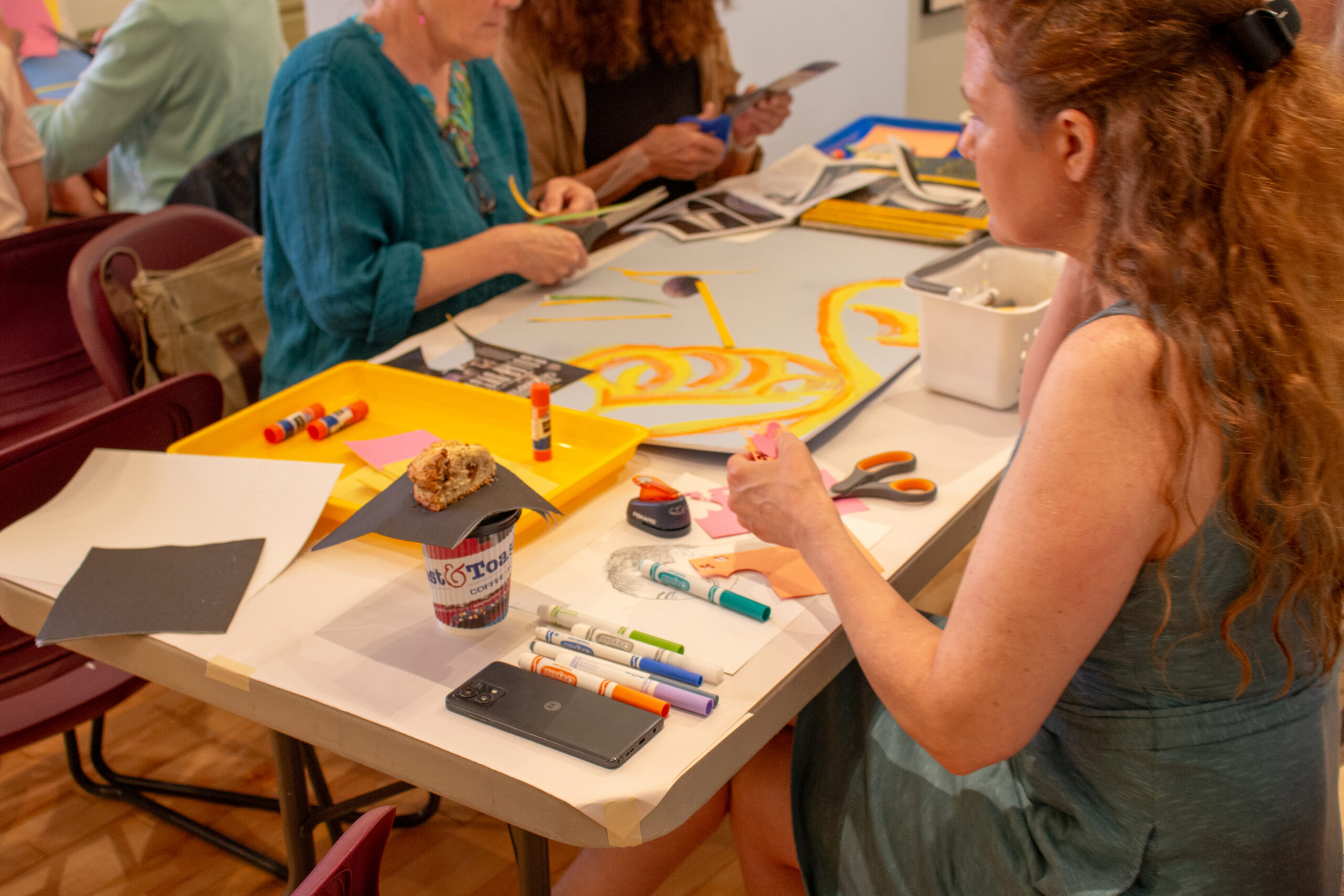 Three people sitting around a table covered with various crafting supplies, painting on a large piece of posterboard.