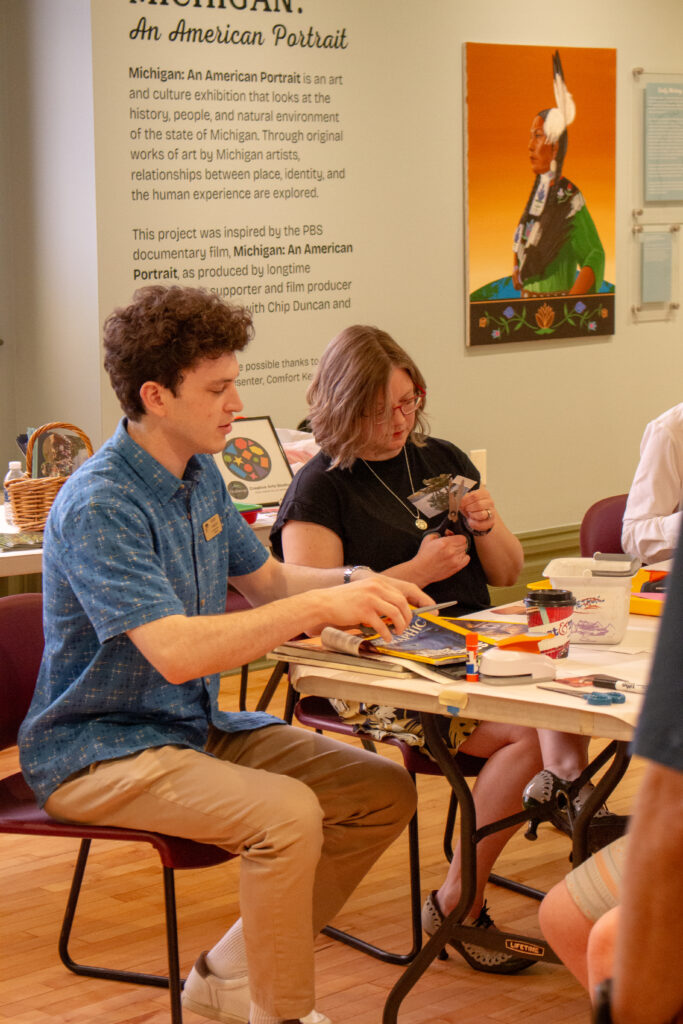 Two people sitting at the corner of a folding table cutting up magazine pages.