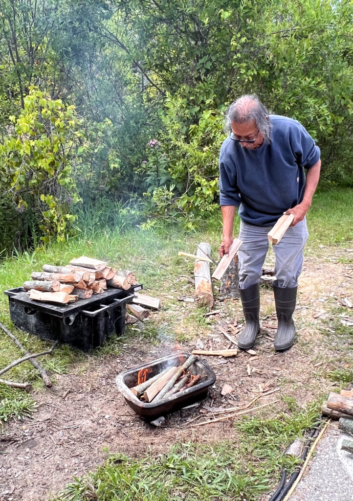 A person wearing a blue long-sleeve shirt, grey pants, and black, knee-high rubber boots holds pieces of chopped wood and throws them into a small fire in a pan in front of him.