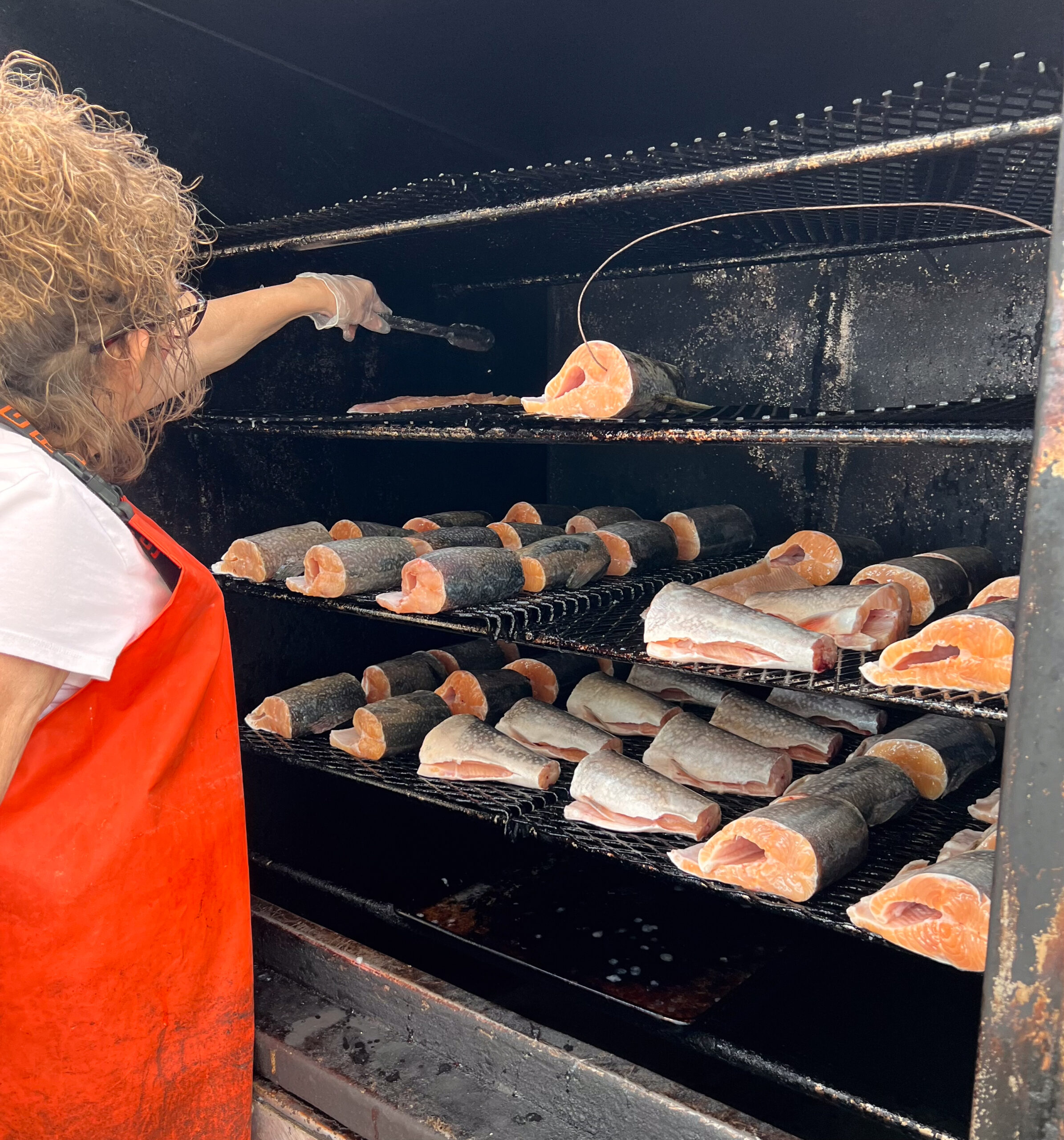 A person in an orange rubber apron using a pair of metal tongs to turn over chinks of fish in a large smoker.