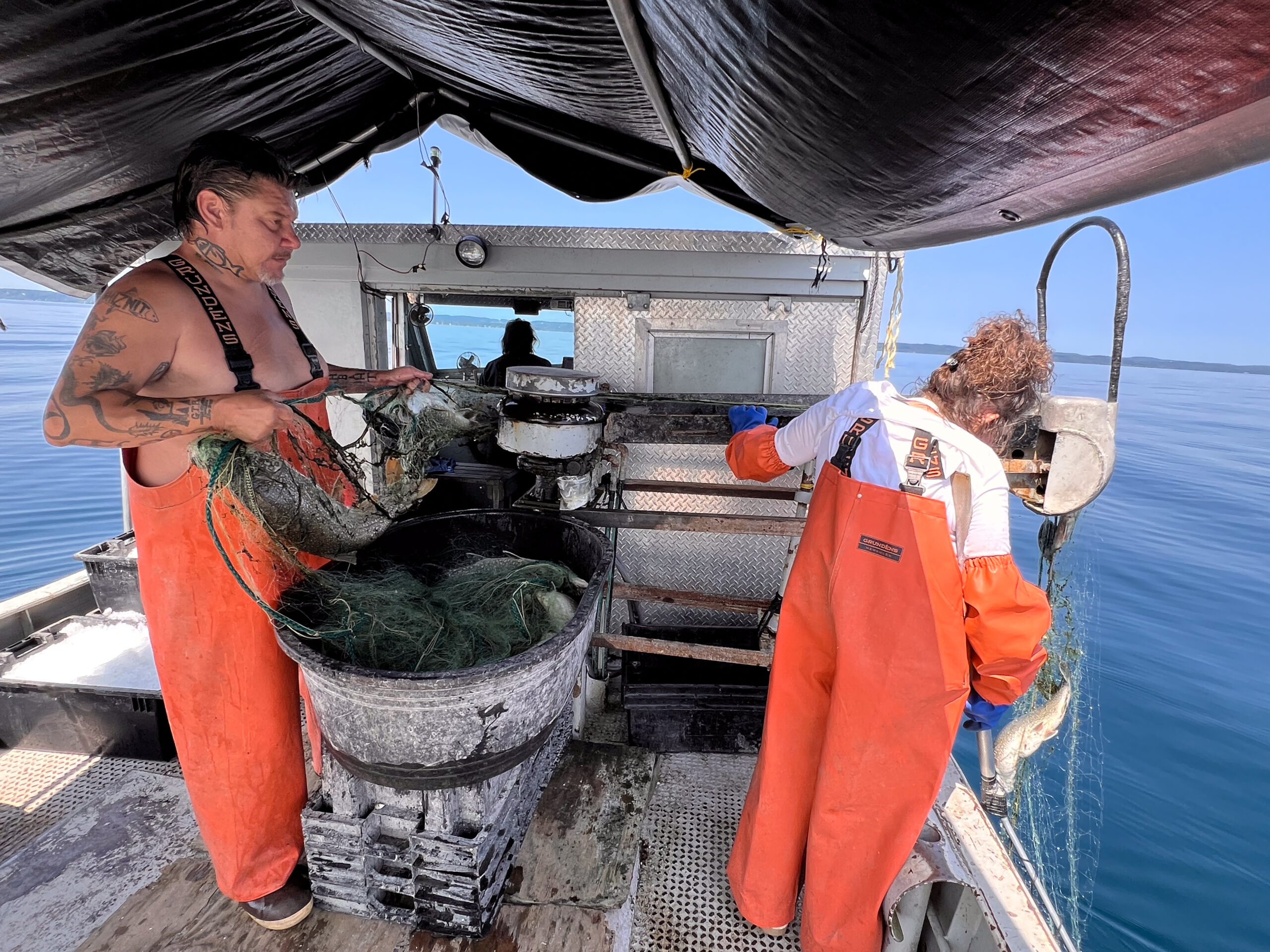 Two people wearing orange rubber overalls on a boat going through a net of fish.