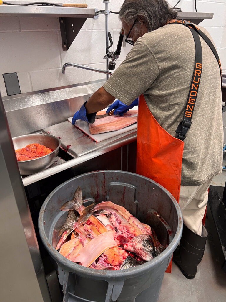 A person wearing an orange rubber apron in a large kitchen filleting fish, with a trash can full of fish carcasses next to him.