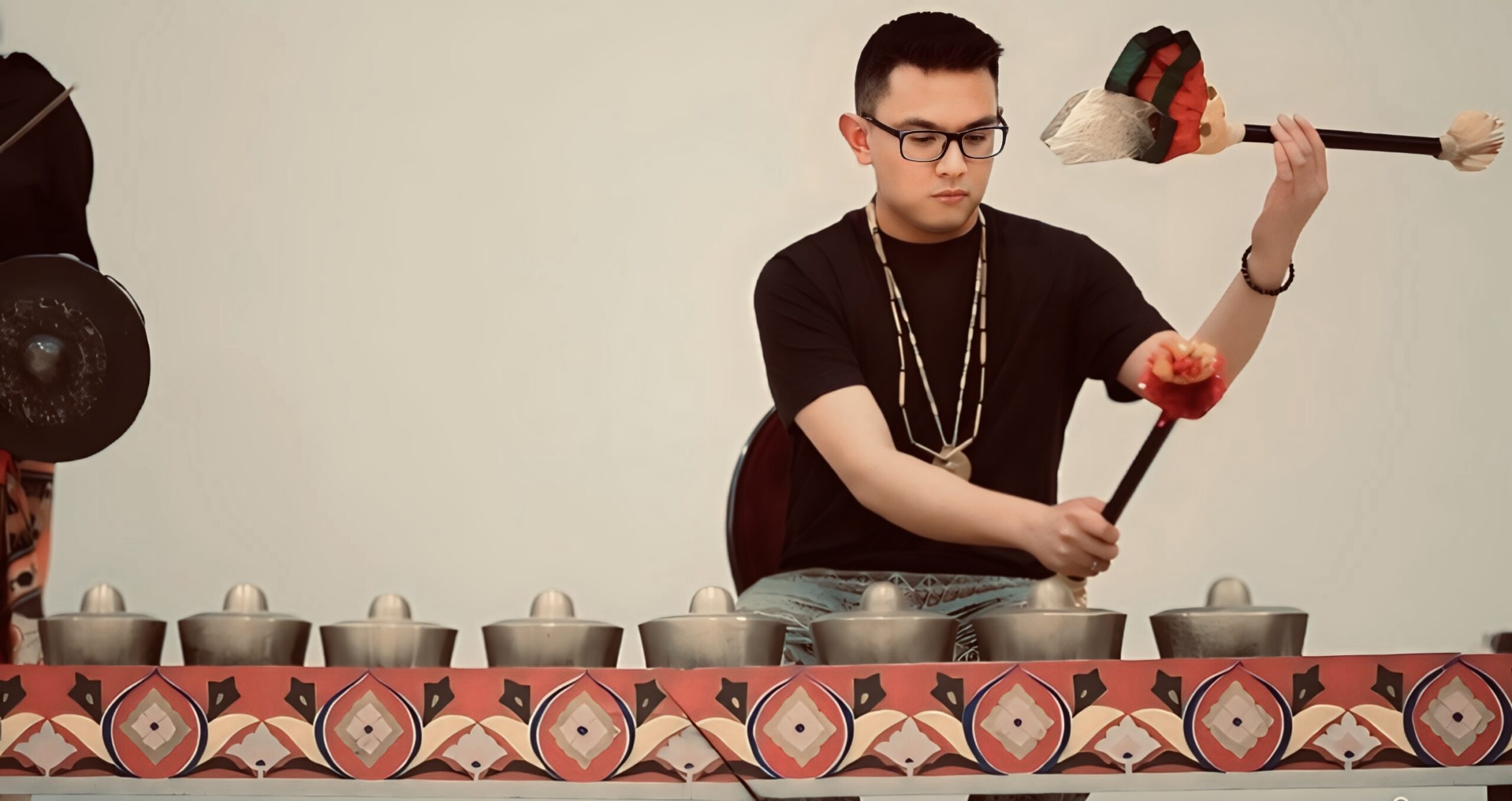 A person with short dark hair and glasses holding two beater sticks, playing the kulintang, an instrument made up of a row of 8 brass gongs suspended on a rope-strung wooden stand.