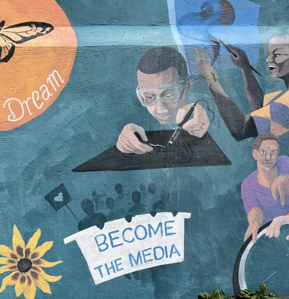 In the center of a section of a wall mural, a young African-American man soldering a computer chip. Below him, a group of people carry a banner that reads, ""Become the Media."" A yellow flower blooms in the lower left hand corner and more people create art in the background.