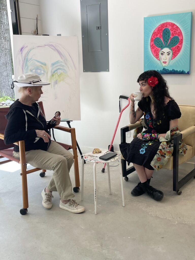 Two people sitting in chairs in a gallery space with an unfinished and finished painting behind them, while a pink cane leans against the wall behind them.