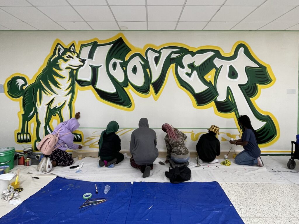 A group of students face away from the camera as they paint a colorful yellow and green mural of a wolf and the word Hoover