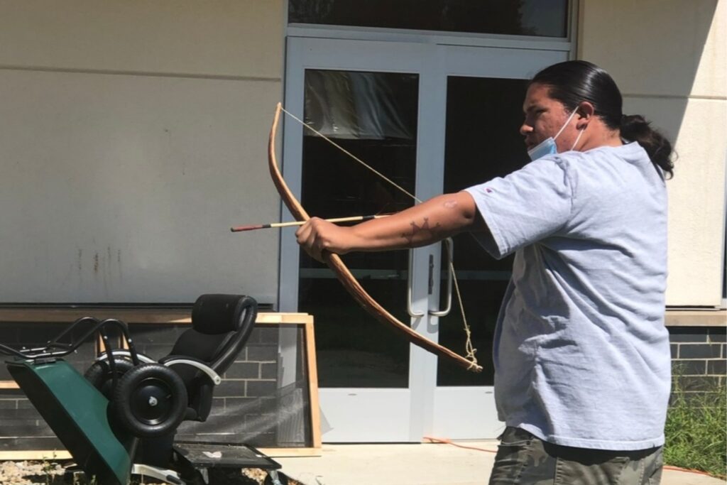 A man with long black hair pulled into a low ponytail aiming a bow and arrow.