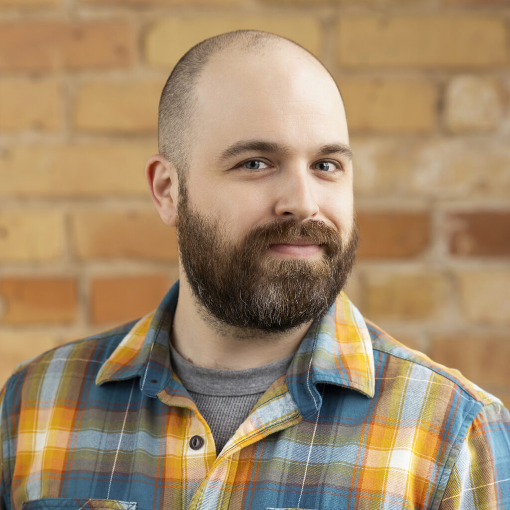 Headshot of a smiling person of light skin tone, with a shaved head and full beard, wearing a plaid shirt.