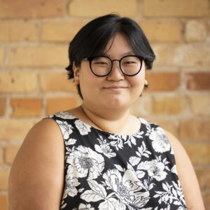 Headshot of a smiling person of medium skin tone, with black hair and glasses, wearing a patterned black floral top