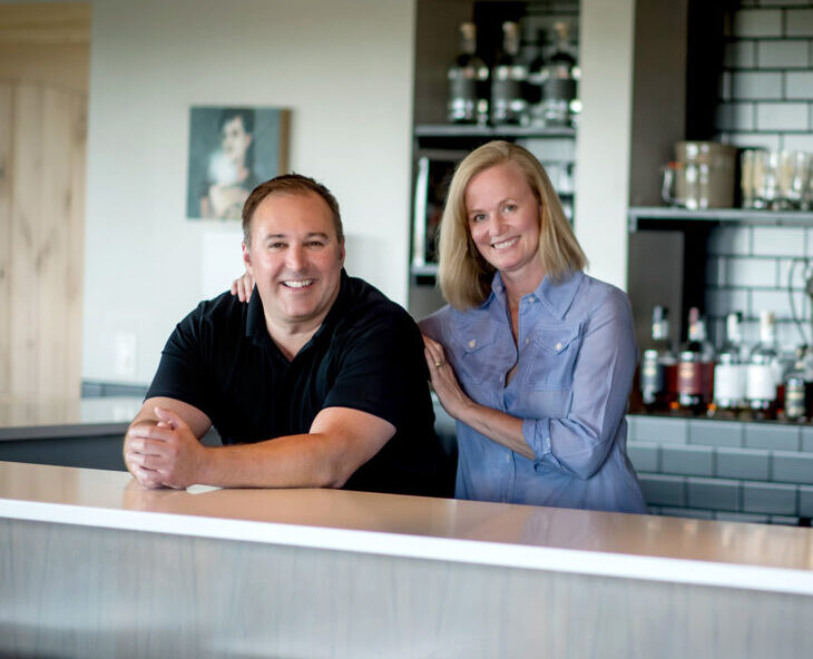 Two people of light skin tone smiling as they stand behind a bar top. One has short dark hair and is wearing a black tshirt, while the other has their arms resting on the person next to them. They have shoulder-length blonde hair and are wearing a light blue button-up shirt.